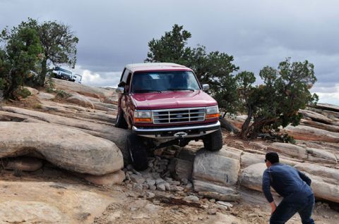 1995 Ford Bronco - A red 1990s Ford Bronco is navigating a rocky, off-road trail. The vehicle is angled up a steep incline, with its tires on large rocks, whi…