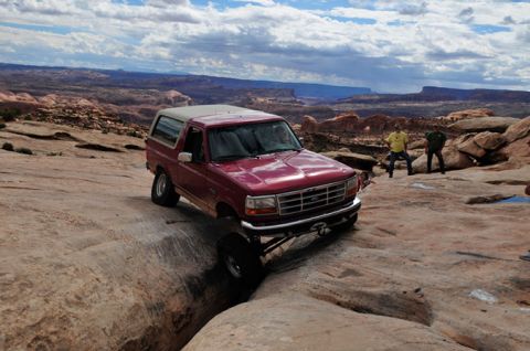 1995 Ford Bronco - A red 1995 Ford Bronco is navigating a rocky, off-road trail with a deep crevice. Two people stand on a rock ledge in the background, obser…