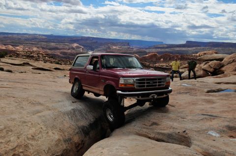 1995 Ford Bronco - A lifted, maroon 1995 Ford Bronco is navigating a rocky, off-road trail with a deep crevice in front of it. Two people stand in the backgro…