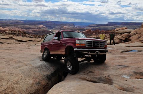 1995 Ford Bronco - A lifted, maroon Ford Bronco is navigating a rocky, off-road trail with a canyon landscape in the background. Two people stand on the rocks…