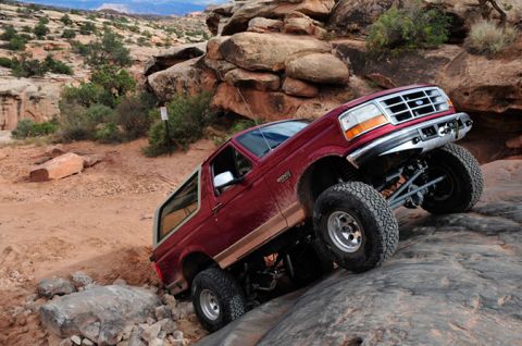 1995 Ford Bronco - A red 1995 Ford Bronco is shown navigating a challenging rocky off-road trail. The vehicle is angled, with its front right wheel high on a…