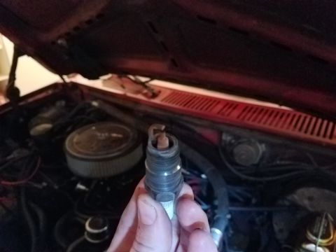 1985 Ford Bronco - A hand holds a spark plug in front of the engine bay of a vehicle with the hood open. The engine features an air filter housing and various…