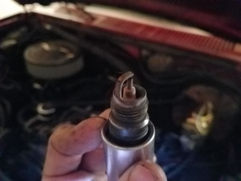 1985 Ford Bronco - A close-up view of a used spark plug held in front of a red vehicle's engine bay. The spark plug shows signs of wear and carbon buildup.