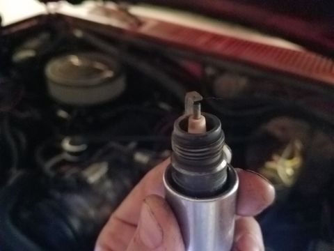 1985 Ford Bronco - A hand holds a spark plug in front of a red vehicle's engine bay. The spark plug appears to have a bent electrode.