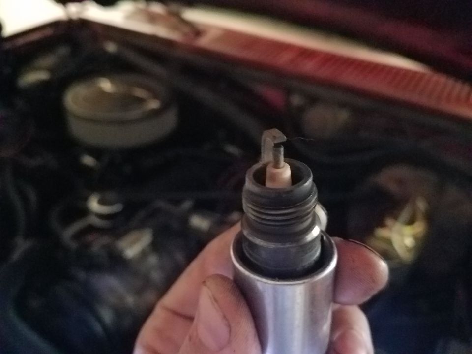 Spark plug with bent electrode held in front of a vehicle engine bay