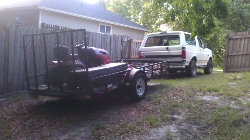 White Ford Bronco parked next to a trailer with a riding lawn mower on it.