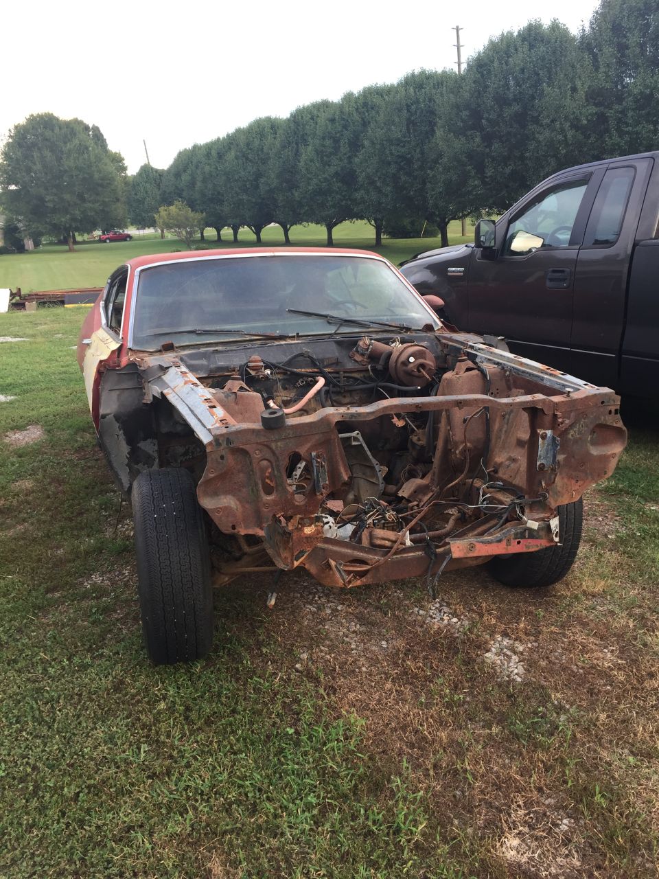 Front view of a rusty 1970 Ford Torino with no hood, parked on grass.