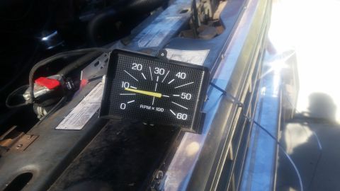 1985 Ford Bronco - A tachometer gauge is shown resting on the engine bay of a vehicle. The gauge reads approximately 1000 RPM, indicated by a yellow needle po…