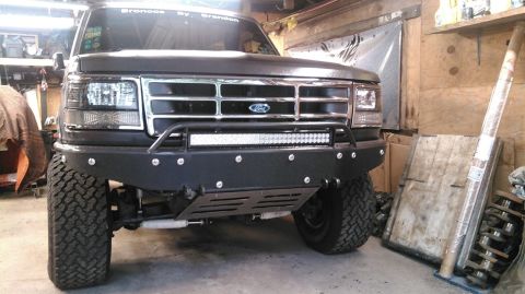 1992 Ford Bronco - Front view of a dark-colored Ford Bronco with a custom black bumper, a light bar, and large off-road tires. Text on the windshield reads "B…