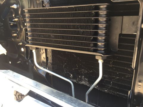 1978 Ford Bronco - A close-up view of a transmission cooler mounted in front of a vehicle's radiator. The cooler has multiple fins and two silver metal lines…