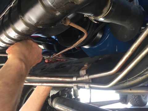 1978 Ford Bronco - A mechanic's hands are visible, using a wrench to work on a metal line attached to the underside of a vehicle. The engine components, inclu…