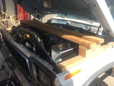 1978 Ford Bronco - The open hood of a white Ford Bronco reveals the engine bay, with several pieces of lumber stacked on top of the fender and radiator suppor…