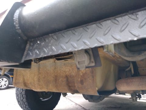 1996 Ford Bronco - A low-angle shot of the underside of a vehicle, showing a rusty fuel tank, exhaust pipe, and a trailer hitch electrical connector. A tire a…
