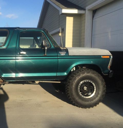 1979 Ford Bronco - The side profile of a lifted, dark green 1979 Ford Bronco is shown parked in front of a garage. The vehicle features large, aggressive tire…
