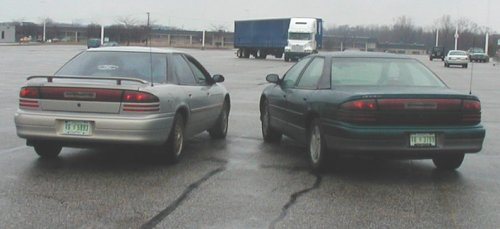 Two sedans parked in a lot, one silver and one dark green. A semi-truck is in the background