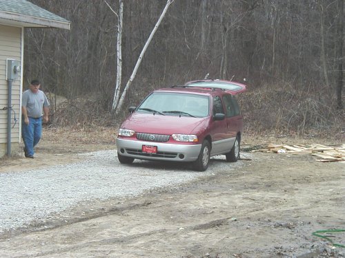 Red minivan with gray lower cladding parked on gravel drive at rural build site