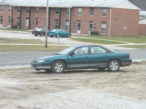 Teal Dodge Intrepid sedan on dirt lot, front 3/4 view, brick apartments behind