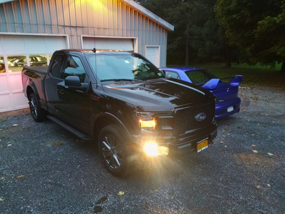 Black Ford F-150 pickup truck parked in front of a garage, with a purple sports car behind it.