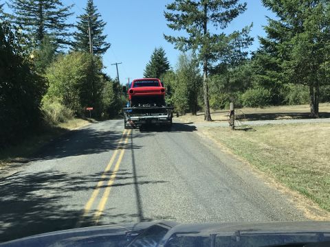 1964 Ford Fairlane - A red classic car is being transported on a flatbed tow truck on a rural road. The car is viewed from behind, showing its rear end and tail…