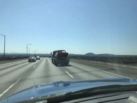 1964 Ford Fairlane - A red classic car is being transported on a flatbed tow truck on a sunny day on a highway. Several other cars are visible on the road ahead.