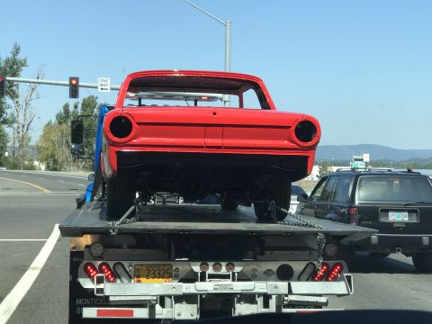 1964 Ford Fairlane - A red car body is being transported on a flatbed tow truck, with a black Jeep visible behind it on the road. The red car appears to be a cl…