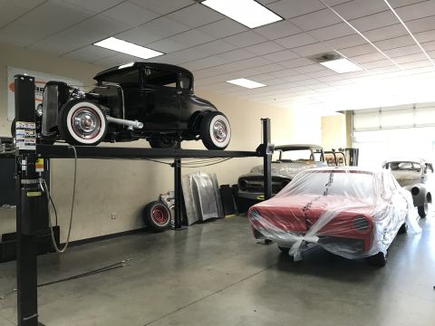 1964 Ford Fairlane - A black 1930s coupe hot rod is elevated on a car lift, while a red classic car covered in plastic wrap is parked in front of it. Several ot…
