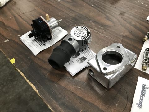 1964 Ford Fairlane - Three automotive parts are displayed on a wooden surface. From left to right, a black and silver fuel pressure regulator, a silver and blac…