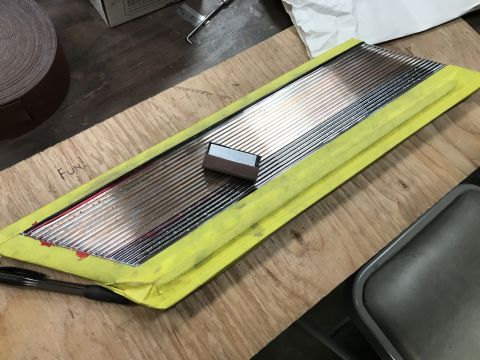 1964 Ford Fairlane - A close-up view shows a chrome trim piece with horizontal ridges, taped down with yellow masking tape on a wooden surface. A small, rectang…