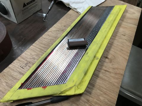 1964 Ford Fairlane - A close-up view shows a chrome trim piece with vertical grooves, taped off with yellow painter's tape. A small rectangular block is placed…