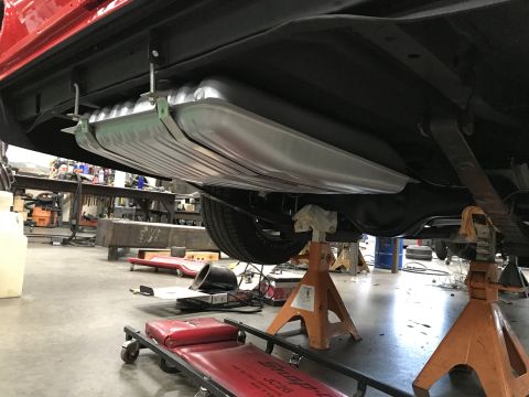 1964 Ford Fairlane - The underside of a classic car shows a new, silver fuel tank being installed, suspended by straps and bolts. The vehicle is raised on orang…
