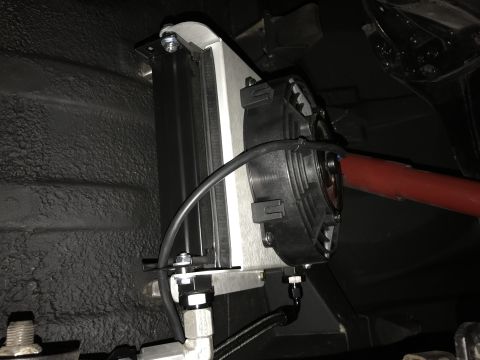 1964 Ford Fairlane - A black electric fan is mounted to a silver oil cooler, which is attached to the textured black undercarriage of a vehicle. A red bar is vi…
