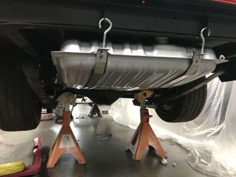 1964 Ford Fairlane - The underside of a vehicle is shown, with a new fuel tank suspended by hooks and straps, supported by jack stands. The rear suspension and…