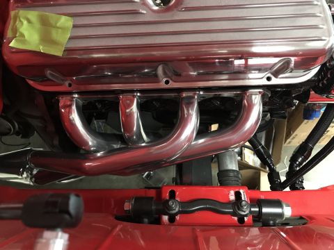 1964 Ford Fairlane - A close-up view of a chrome exhaust manifold and silver valve cover on a V8 engine, with red painted components in the foreground. Yellow t…