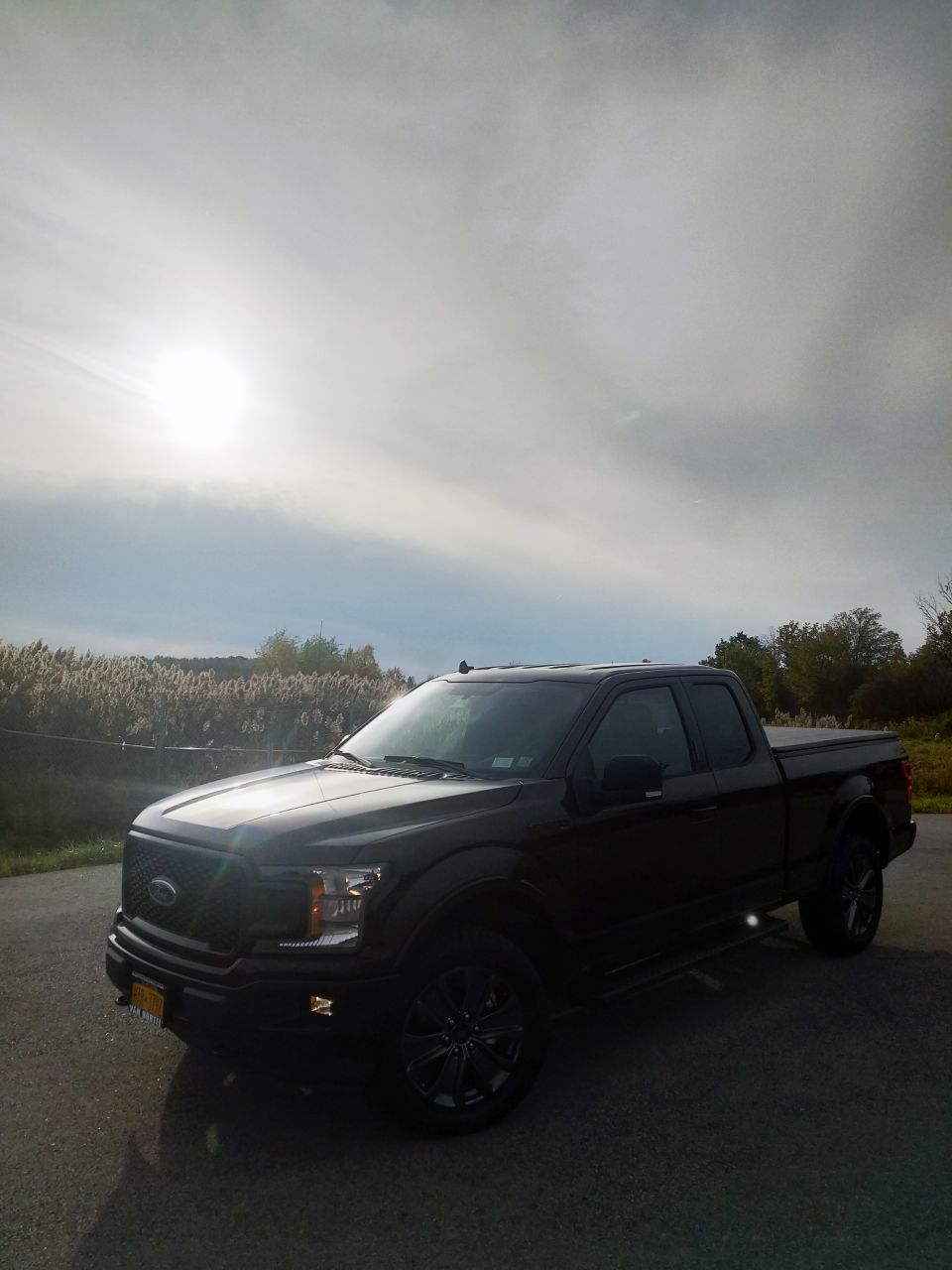 Black 2018 Ford F-150 pickup truck parked on asphalt, with a field and trees in the background under a cloudy sky with the sun visible