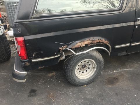 1992 Ford Bronco - The rear quarter panel and wheel well of a black Ford Bronco are heavily rusted and damaged. The vehicle is parked outdoors on an asphalt s…