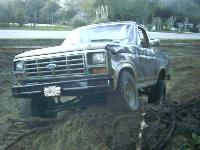 Damaged topless 1980s Ford Bronco with crushed roof sitting in a muddy root-filled pit