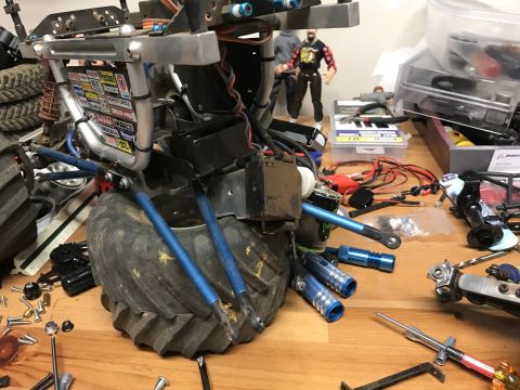 1989 Ford Bronco - A partially assembled remote-control monster truck chassis is shown on a wooden workbench, surrounded by tools and spare parts. The chassis…