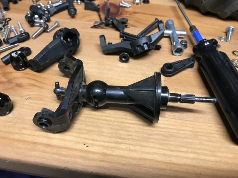 1989 Ford Bronco - A collection of black plastic and metal RC car parts are scattered on a wooden surface. Visible parts include gears, suspension components,…