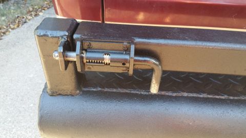 1996 Ford Bronco - A close-up view of a black metal latch mechanism mounted on the side of a vehicle's bumper. The latch has a spring-loaded bolt with a bent…