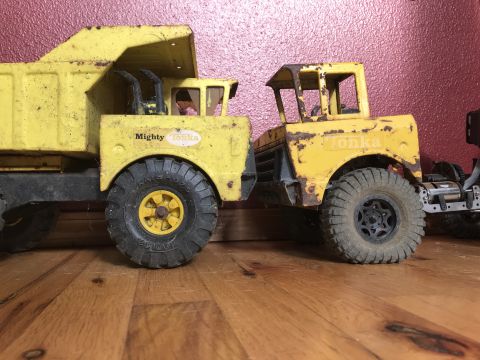 1989 Ford Bronco - Two vintage yellow Tonka toy trucks are positioned side-by-side on a wooden floor in front of a textured pink wall. The truck on the left i…