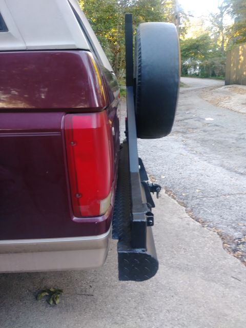 1996 Ford Bronco - The rear passenger side of a dark red Ford Bronco with a tan lower trim and a black swing-away spare tire carrier mounted to the rear bumpe…