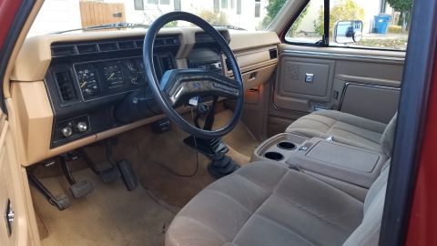 1985 Ford Bronco - The interior of a red Ford Bronco is shown, featuring tan upholstery, a tan dashboard with gauges, and a black steering wheel. The view inc…