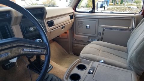1985 Ford Bronco - The interior of a vintage Ford Bronco is shown, featuring a tan dashboard, steering wheel, and cloth seat. The center console with cup hold…