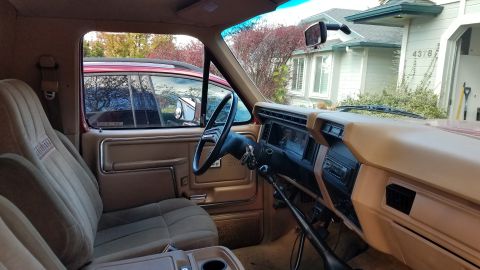 1985 Ford Bronco - Interior shot of a tan-trimmed Ford truck cab showing the dash, steering wheel and cloth seat, looking out through the windshield at a hous…