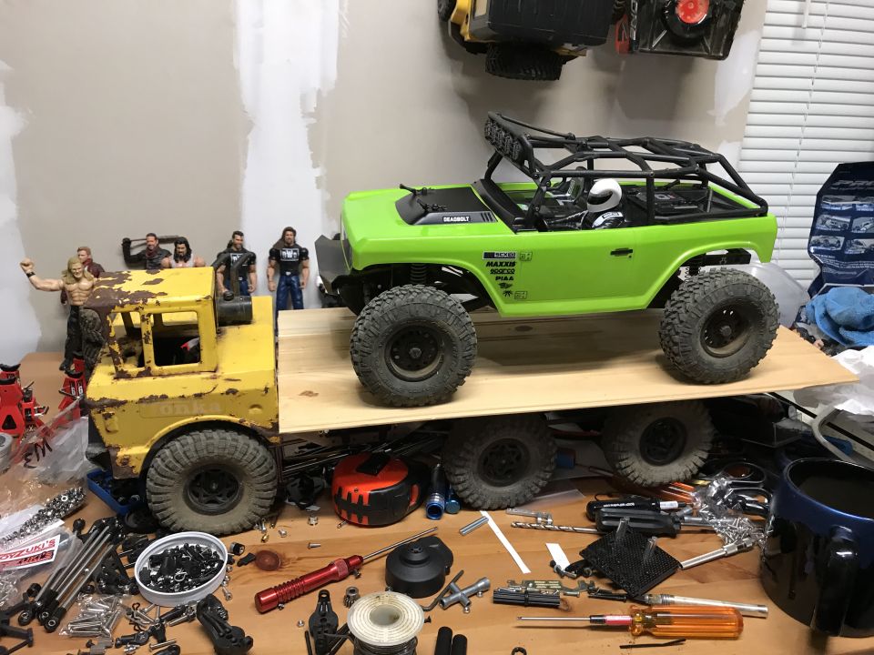 Green RC rock crawler on a wooden platform above a yellow Tonka truck, surrounded by tools and parts.