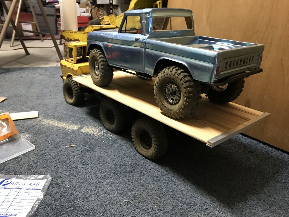 Blue Chevrolet pickup truck on a wooden trailer towed by yellow heavy equipment.