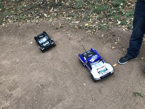 1989 Ford Bronco - Two remote control trucks are parked on a dirt path, with a person's legs visible on the right side of the frame.