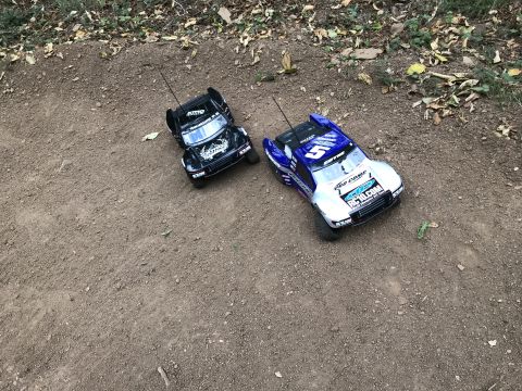 1989 Ford Bronco - Two remote control trucks, one black and one blue and silver, are positioned on a dirt track.