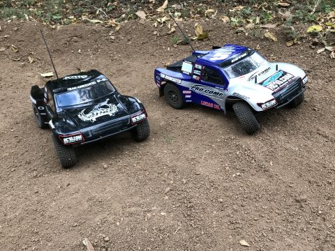 1989 Ford Bronco - Two RC trucks, a black Team Associated and a blue/silver Pro Comp, sit on a dirt track.
