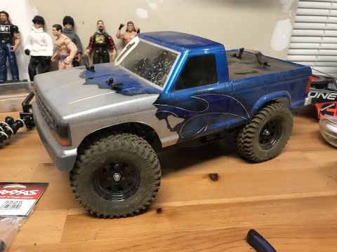 1989 Ford Bronco - A remote-controlled truck with custom flame decals sits on a wooden surface, with action figures visible behind it.